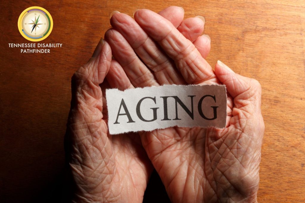 Elderly hands open and resting on a wooden surface. Hands are holding a piece of paper with the word AGING typed in all capital letters. Pathfinder branding graphic with colorful compass and Tennessee Disability Pathfinder underneath in white font.